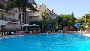 Une piscine entourée de chaises longues et de parasols, elle-même bordée d’un immeuble à plusieurs étages doté de balcons.