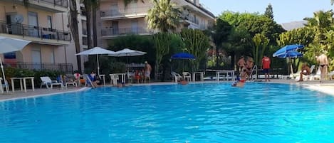 A swimming pool with lounge chairs and umbrellas, surrounded by a multi-story building with balconies.