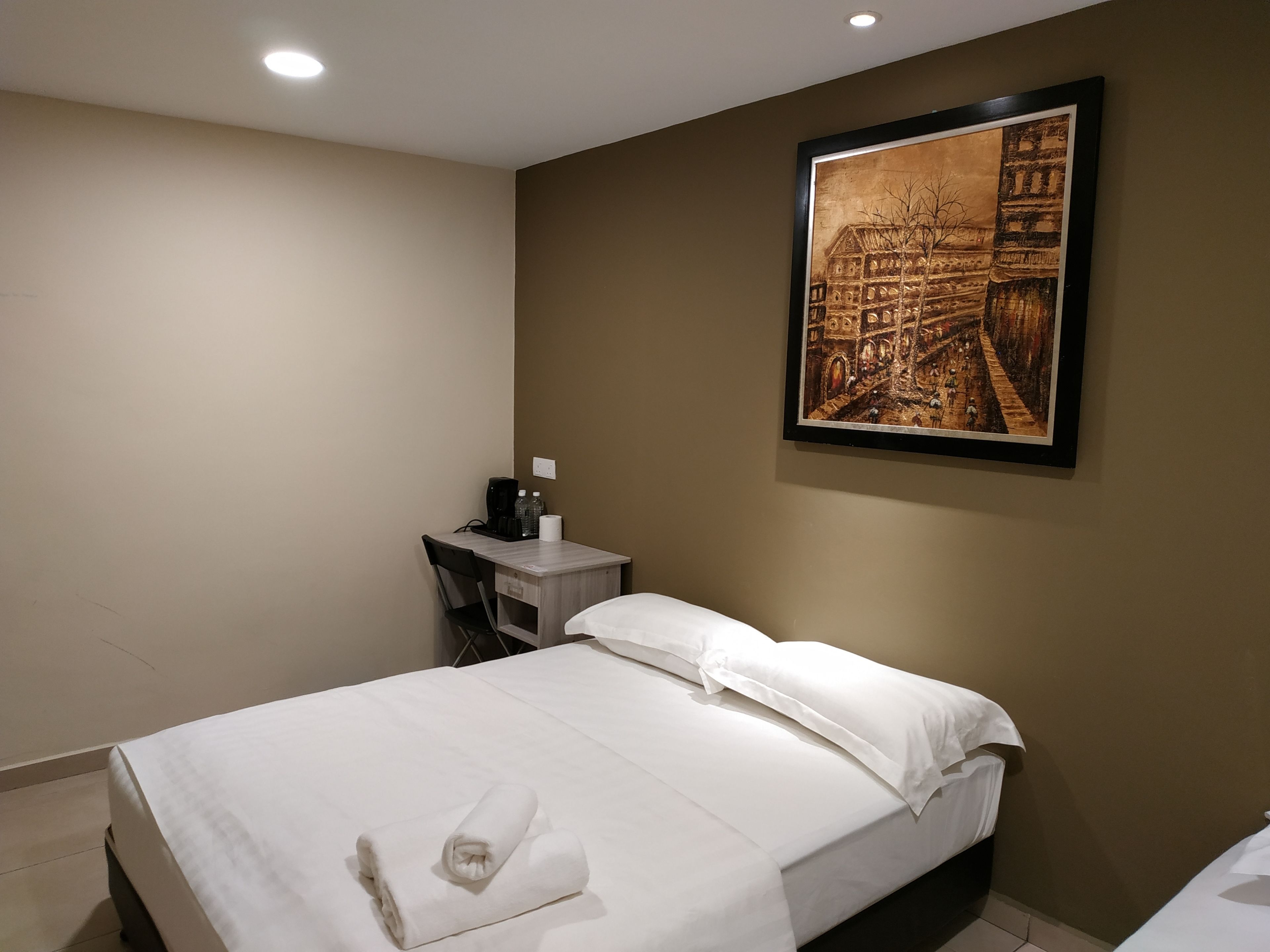 A hotel room with a bed, a desk, a chair, a painting on the wall, and a coffee machine.