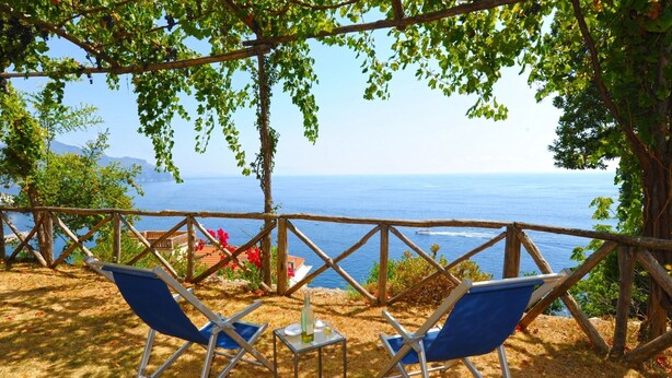 Eine Terrasse mit zwei blauen Stühlen, einem Tisch mit einer Flasche und Blick auf das Meer.