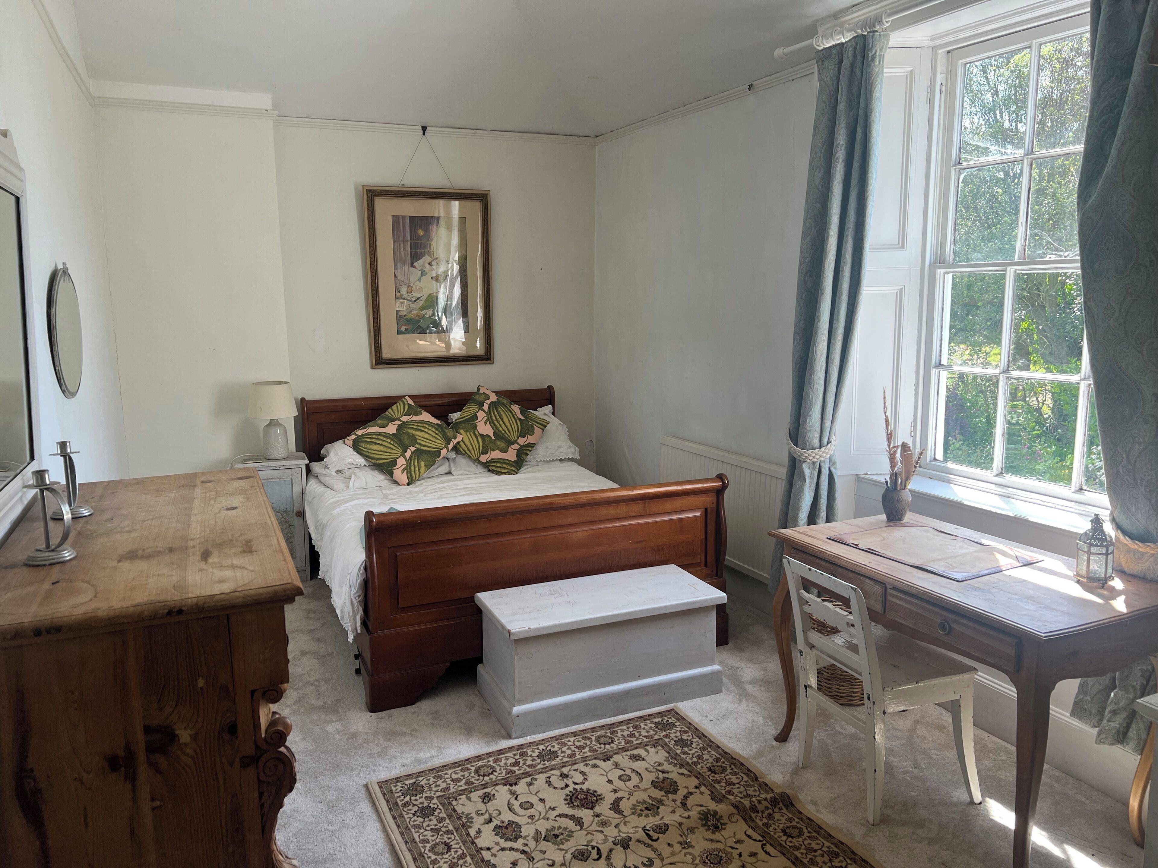 A bedroom with a wooden bed, a bedside table, a desk, a chair, a rug, a window with curtains, and a framed picture on the wall.