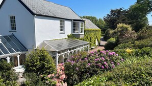 Une maison blanche dotée d’une véranda, entourée d’une végétation luxuriante et de plantes en fleurs.