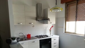 Une cuisine avec des meubles blancs, une cuisinière à gaz, un évier et une fenêtre avec des stores.