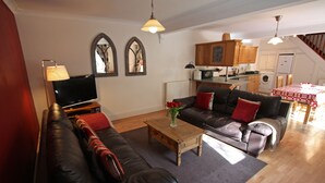 A living room with a sofa, coffee table, and a television. There is a kitchen area with cabinets and a refrigerator.