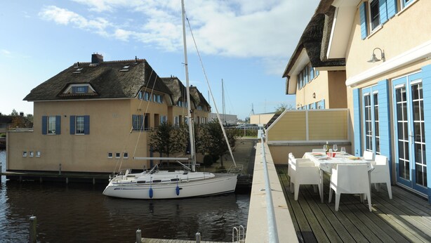 Una propiedad frente al mar con un velero amarrado, un comedor con mesas al aire libre y un edificio con contraventanas azules.