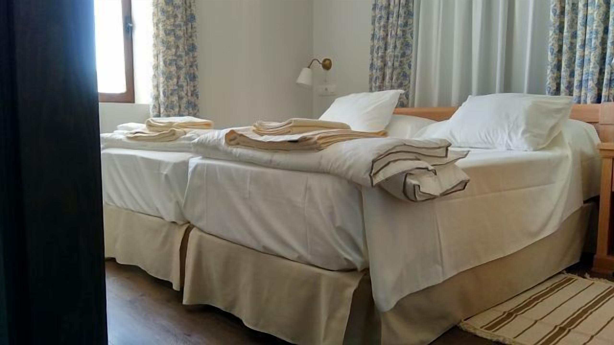 Habitación de hotel con dos camas, una mesita de noche, una lámpara y una ventana con cortinas.