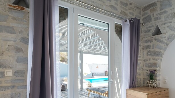 A bedroom with a stone wall, a sliding glass door leading to a pool area, a wooden dresser, and a patterned bedspread. - Paros Nereids Villas Estate with private pools and amazing  view (Paros)