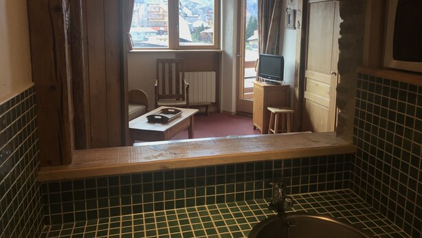 A compact room with a window, a wooden chair, a small table, a television on a cabinet, and a tiled backsplash.