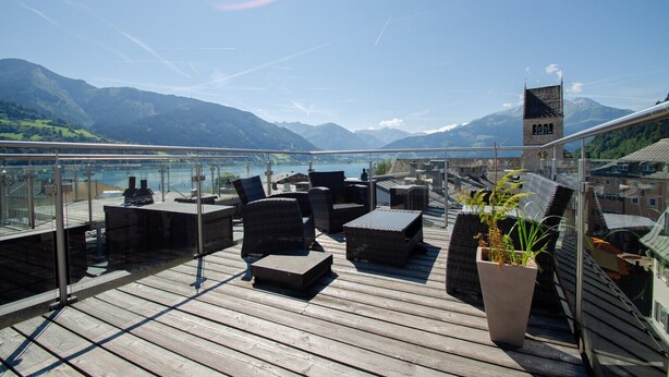 Een houten terras met buitenmeubilair met uitzicht op een meer en bergen.