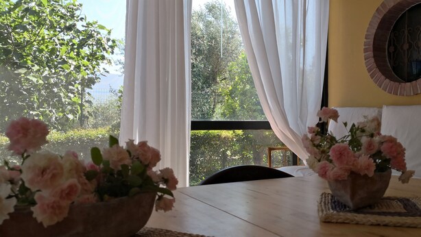 A room with a wooden table, two flower vases, a round mirror, and a view of greenery through a window with white curtains. - Vacation House "La Caprignola" (Buccino)