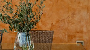 A table with a glass vase containing green foliage, a wicker chair, and a wall with a warm, orange hue.