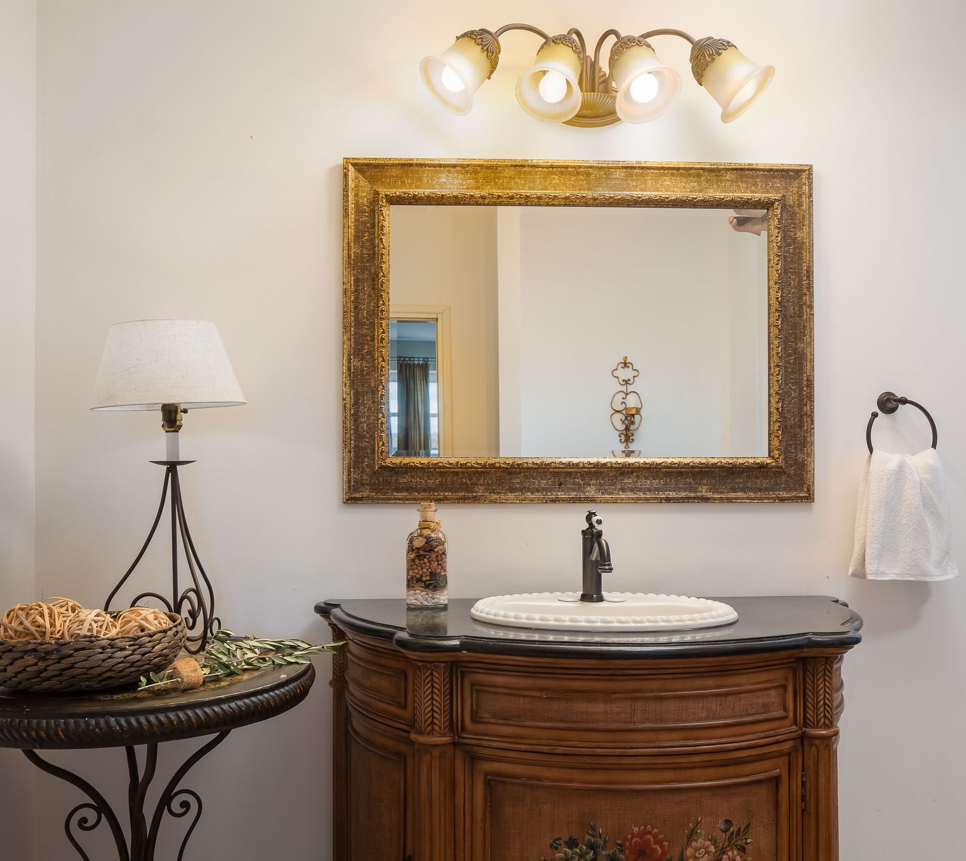 A bathroom with a wooden vanity, a round mirror with a gold frame, a wall-mounted light fixture, a small table with a lamp, and a towel rack.