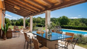 A covered outdoor area with a glass-top table and wicker chairs, overlooking a swimming pool and lush greenery.