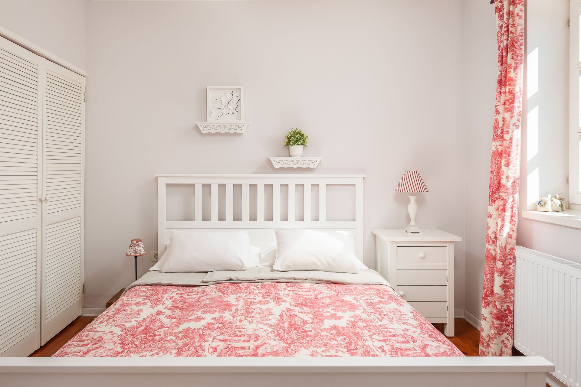 Un dormitorio con una cama blanca, una mesita de noche, un aparador, una ventana con cortinas florales y un radiador blanco.