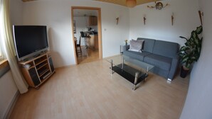 Een woonkamer met een flatscreen televisie op een houten standaard, een grijze bank, een glazen salontafel en een potplant.