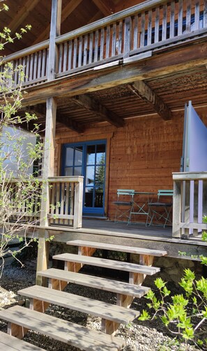 Een houten hutje met een veranda, een blauwe deur en twee opklapbare stoelen.