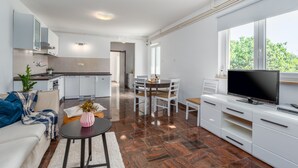Ein modernes Wohnzimmer mit einer Couch, einem Couchtisch und einem Fernseher. Es gibt einen Essbereich mit Tisch und Stühlen. Im Hintergrund ist eine Küche zu sehen.