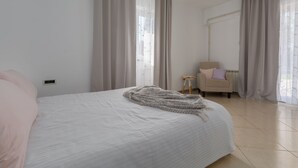Ein ordentlich bezogenes Bett mit weißer Decke und grauer Tagesdecke, ein kleiner runder Tisch mit einem Stuhl und ein großes Fenster mit durchsichtigen Vorhängen.