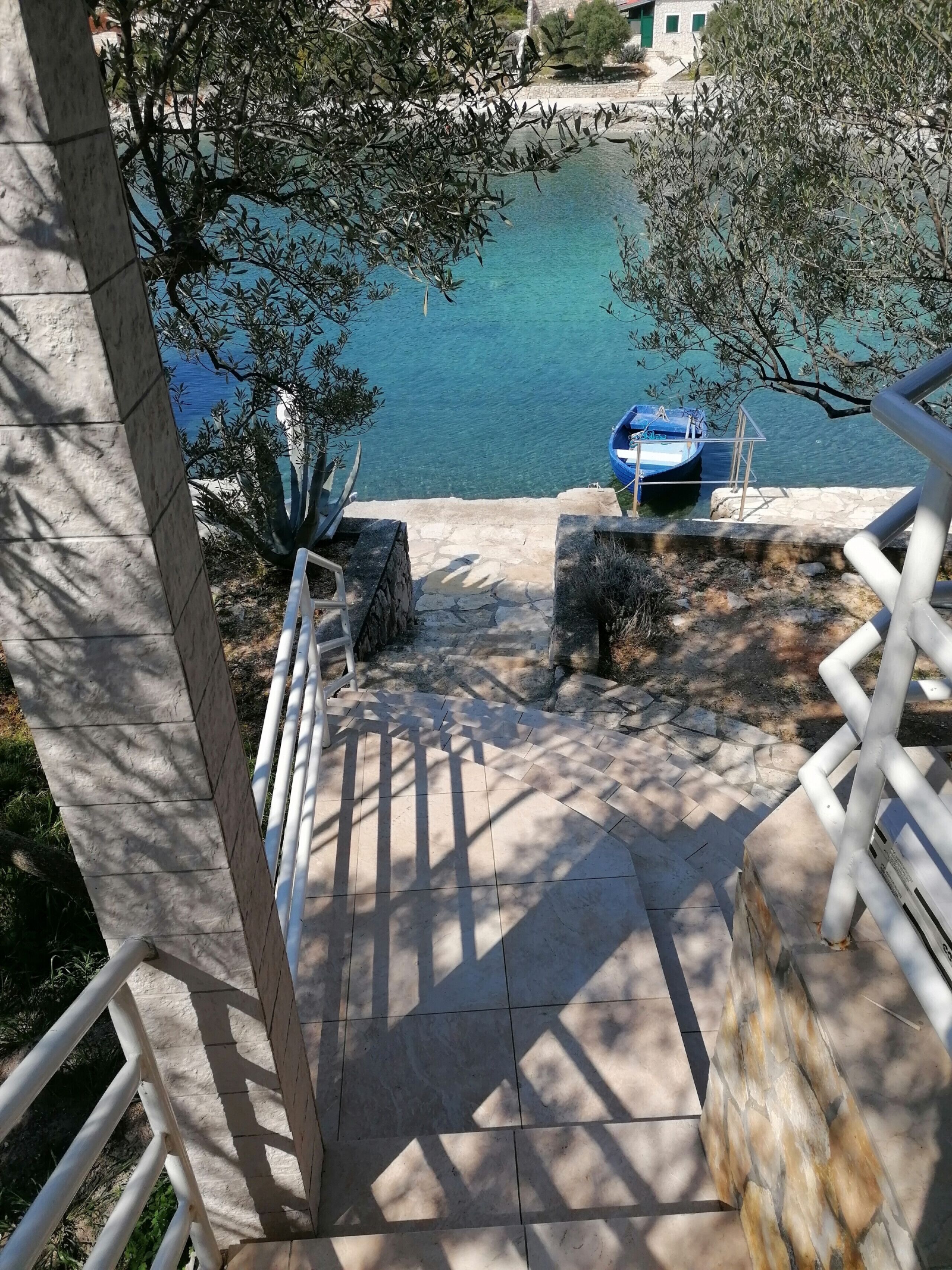 A stone staircase leading to a dock with a blue boat, surrounded by olive trees and a clear view of the water.