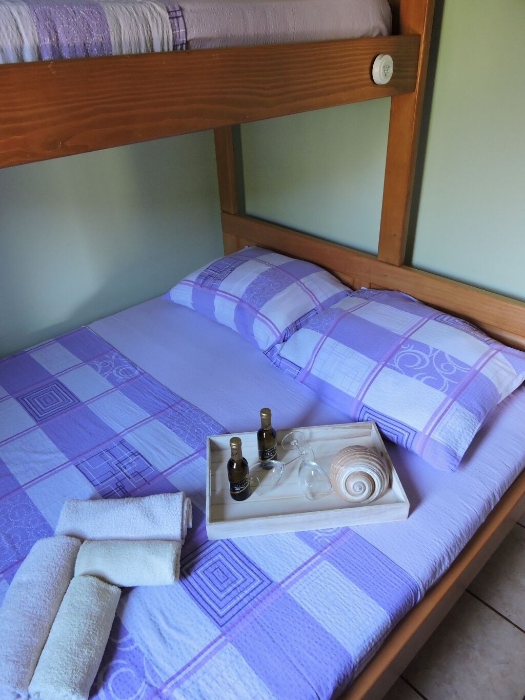 Ein Einzelbett mit hölzernem Gestell, eine violett-weiße karierte Bettdecke, zwei Flaschen, ein Glas und ein zusammenrolltes Handtuch auf dem Bett.