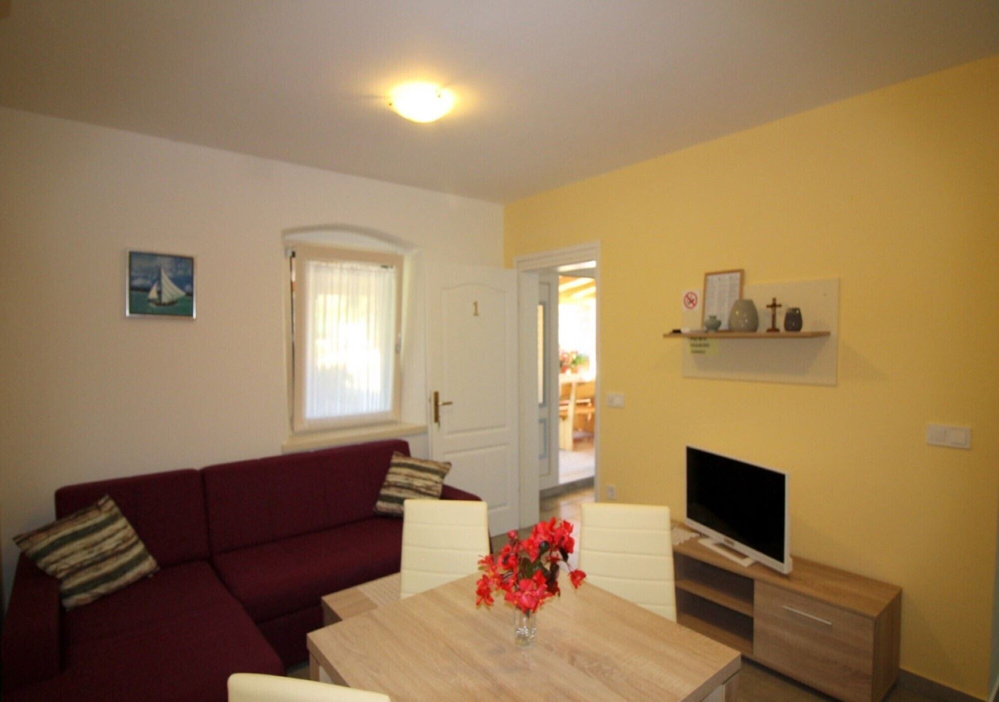 A living room with a red sofa, a wooden dining table with a vase of flowers, a flat-screen TV on a wooden stand, and a door leading to another room.