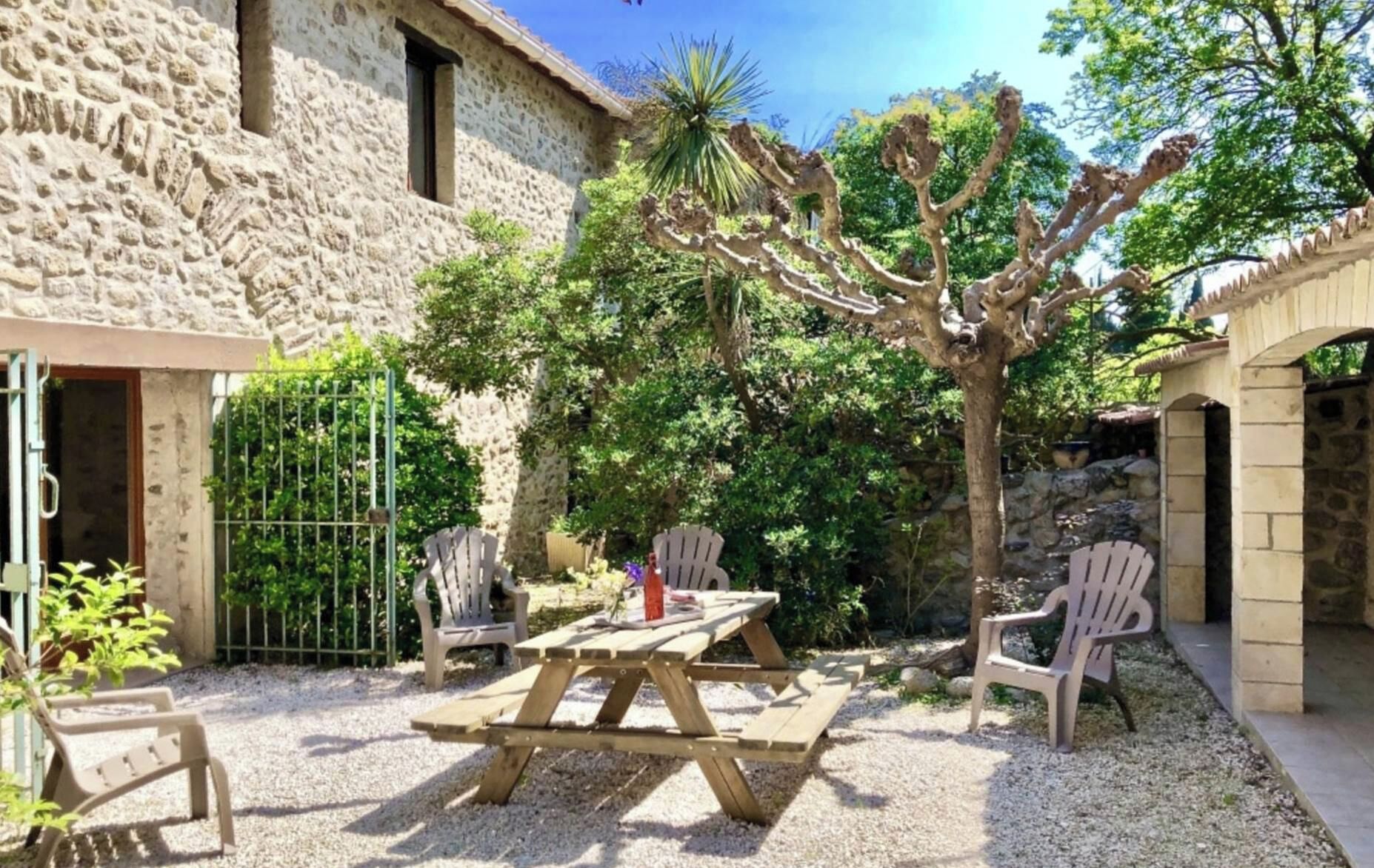 A stone building with a patio area featuring a wooden table and chairs, surrounded by trees and gravel.