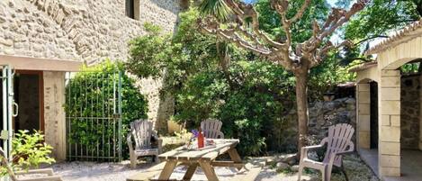 A stone building with a patio area featuring a wooden table and chairs, surrounded by trees and gravel.