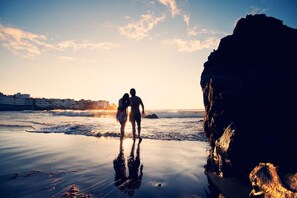 Escena de playa con una gran formación rocosa y una pareja caminando hacia el agua.
