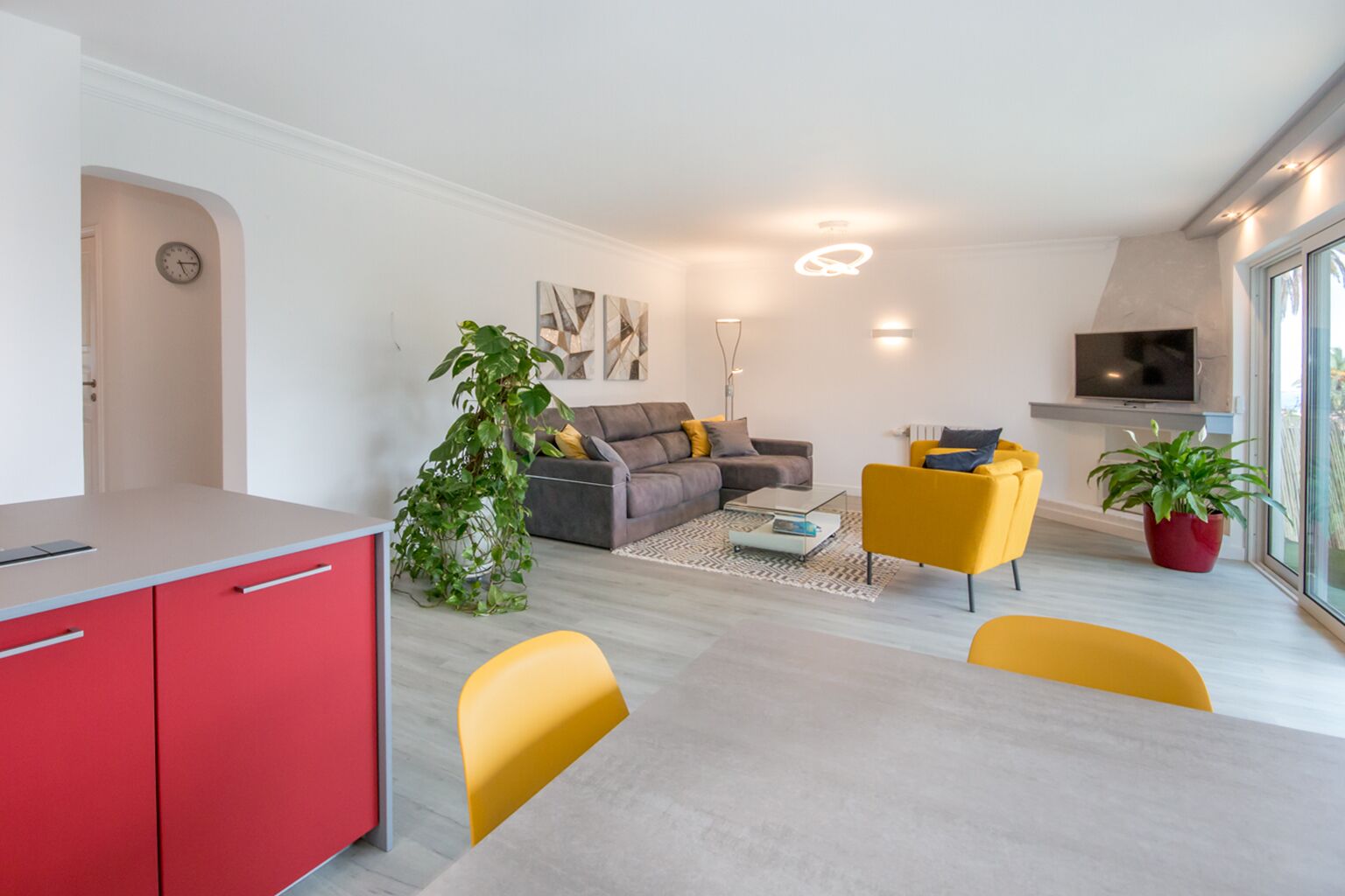 A modern living room with a grey sofa, yellow chairs, a red kitchen counter, and a large window.
