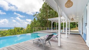 A wooden deck with a pool, lounge chairs, and a covered area with a view of the ocean.