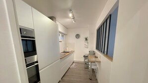 A narrow kitchen with white cabinets, a built-in oven, a sink, and a window with blinds. - Apartment in Deauville center with PARKING and BEACH CABIN (Deauville)