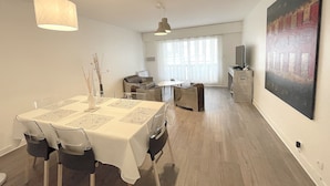 A dining area with a table set for two, a sofa, a TV, and a painting on the wall. - Apartment in Deauville center with PARKING and BEACH CABIN (Deauville)
