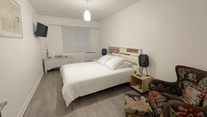 A bedroom with a bed, bedside tables, a chair, a TV, and a window with blinds. - Apartment in Deauville center with PARKING and BEACH CABIN (Deauville)