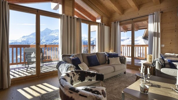 Ein gemütliches Wohnzimmer mit einer Couch, einem Sessel und einem Couchtisch, großen Fenstern mit Vorhängen und Blick auf schneebedeckte Berge.