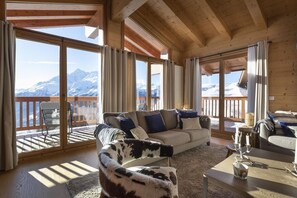 Un salon chaleureux avec un canapé, un fauteuil et une table basse, de grandes fenêtres avec des rideaux et une vue sur des montagnes enneigées.
