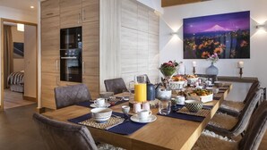 Ein Essbereich mit Holztisch, gedeckt für das Frühstück, eine Küche mit modernen Geräten und ein Gemälde einer Berglandschaft an der Wand.