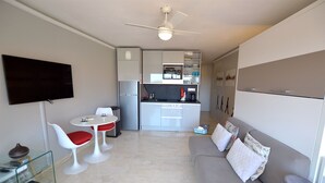 A compact living space with a kitchenette, a dining area with a glass table and red chairs, a grey sofa with decorative pillows, and a flat-screen TV mounted on the wall. - AMAZING APARTMENT LOFT NEAR THE BEACHES (Cap-D'ail)