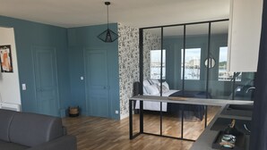 Un espace de vie moderne avec un canapé gris, une petite table ronde, un coin repas séparé par une cloison en verre et une vue sur l’extérieur.