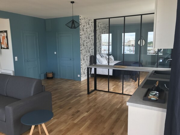 Ett modernt vardagsrum med en grå soffa, ett litet runt bord, en matplats avskild med en glasvägg och utsikt över omgivningen.