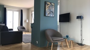 Un salon moderne avec un fauteuil gris, une petite table sur laquelle se trouve une tasse bleue, un lampadaire et un téléviseur à écran plat fixé au mur.
