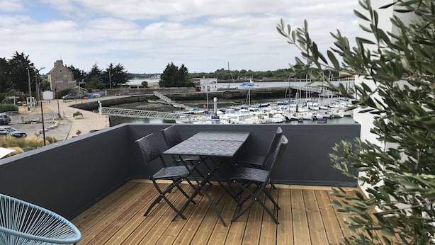 Une terrasse en bois avec une table et des chaises, donnant sur une marina où sont amarrés des bateaux.