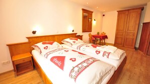 Une chambre avec un lit double recouvert d’une couvre-lit à motifs rouges et blancs, une table de chevet en bois, une armoire en bois et une petite table recouverte d’une nappe rouge. Le sol est en bois et les murs sont blancs.