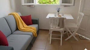 A modern living room with a grey sofa, red and yellow cushions, a white table with a vase and cups, and a white chair. - Haven of peace Cosy air-conditioned studio 16 m2+ Swimming pool for 2 persons (Porto-Vecchio)