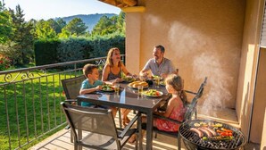 Une famille prend son repas en plein air, sur une terrasse équipée d’un barbecue, avec vue sur un jardin luxuriant.