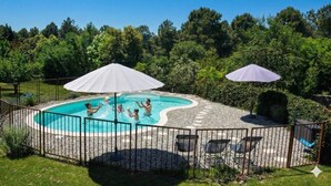 Une piscine avec deux parasols, entourée d’une clôture, et une pelouse avec des arbres en arrière-plan.