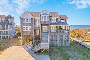 A multi-story beachfront house with a balcony, a staircase leading to the upper floor, and a wooden deck.