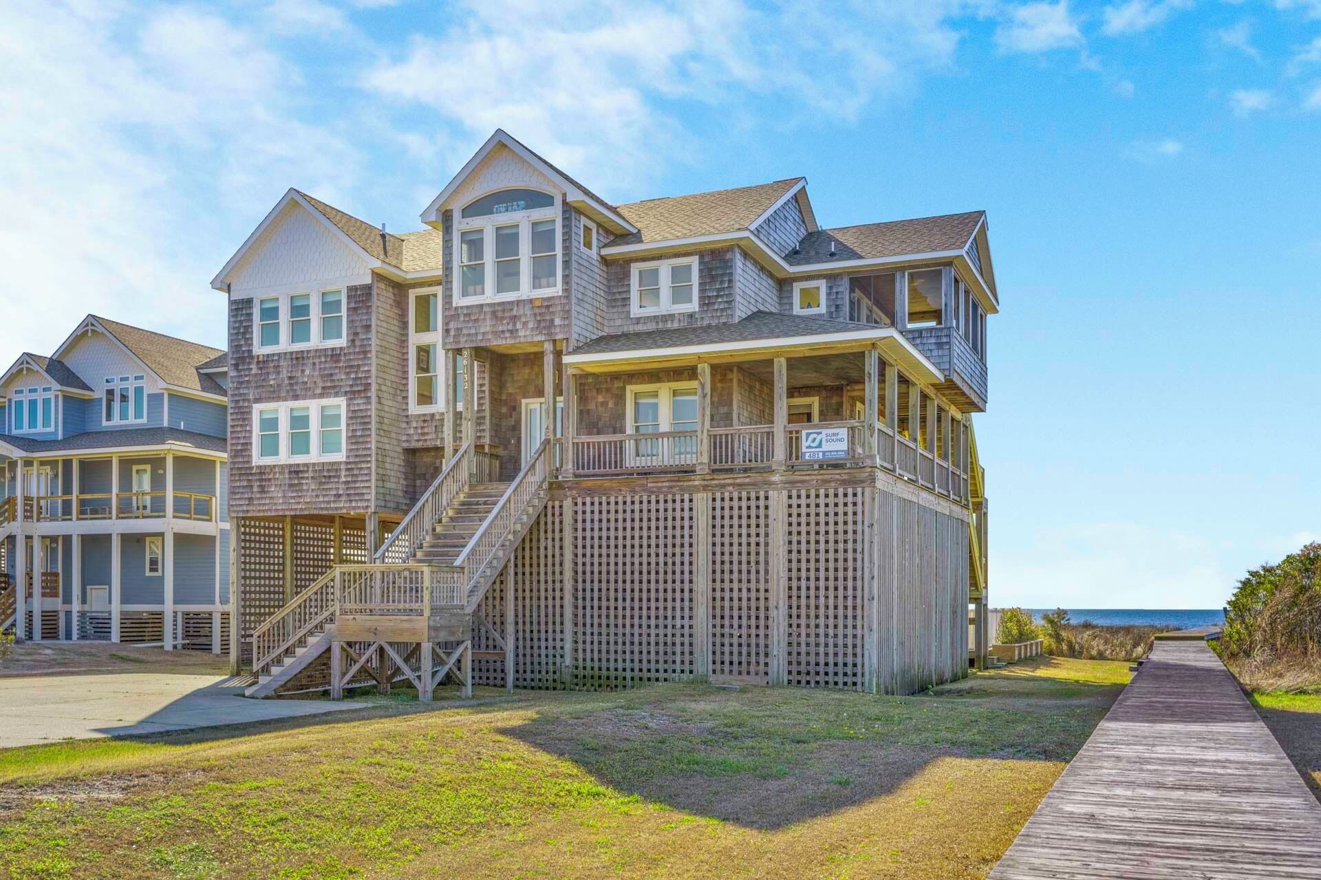 A multi-story beachfront property with a wooden deck, a staircase leading to a balcony, and a view of the ocean.