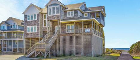 A multi-story beachfront property with a wooden deck, a staircase leading to a balcony, and a view of the ocean.