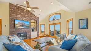 A spacious living room with a fireplace, a ceiling fan, and large windows offering a view of the beach. - Sweet Tranquility – Soundfront Retreat w/ Pool, Hot Tub, Game Room & Tiki Bar (Salvo)