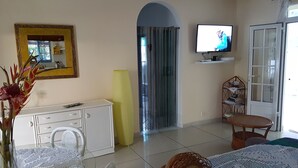 Uma sala de estar com um armário branco, uma televisão fixada na parede, um vaso amarelo, uma cadeira de vime e uma mesa com toalha rendada.